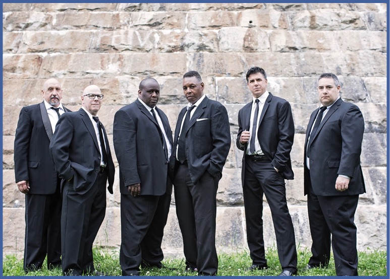 the band stands in black suits in front of a stone wall