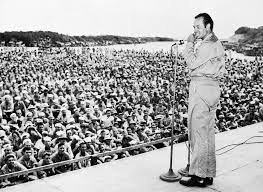 black and white photo of Bob Hope on stage in front of a mic, in front of an enormous crowd of troops 