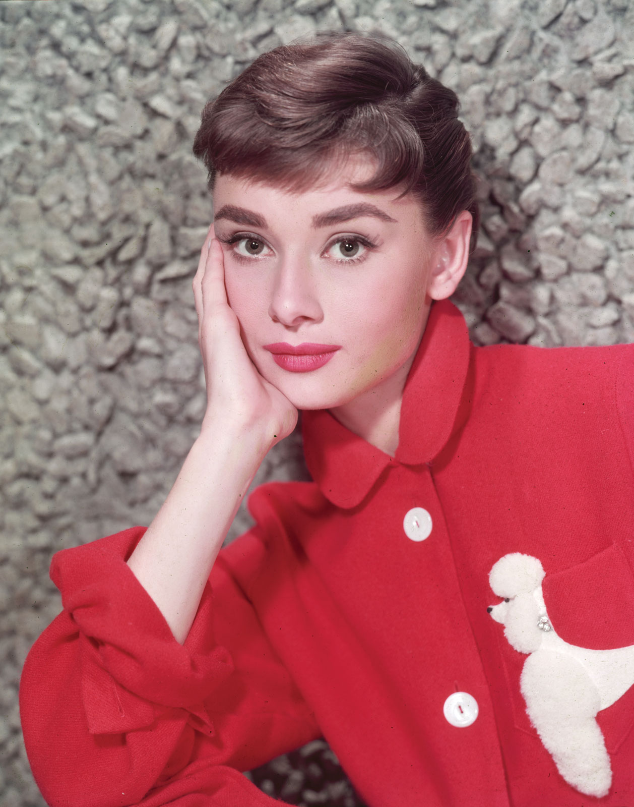 Hepburn in a red blouse with a white poodle decoration leaning on her hand 