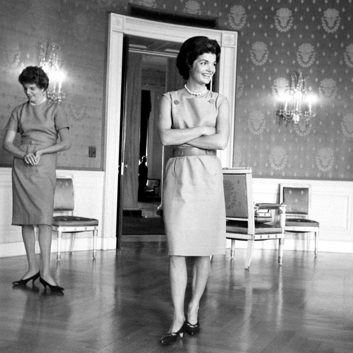 Black and white photo of Jackie Kennedy standing in the center of a room. Behind her is another woman and a few chairs. 