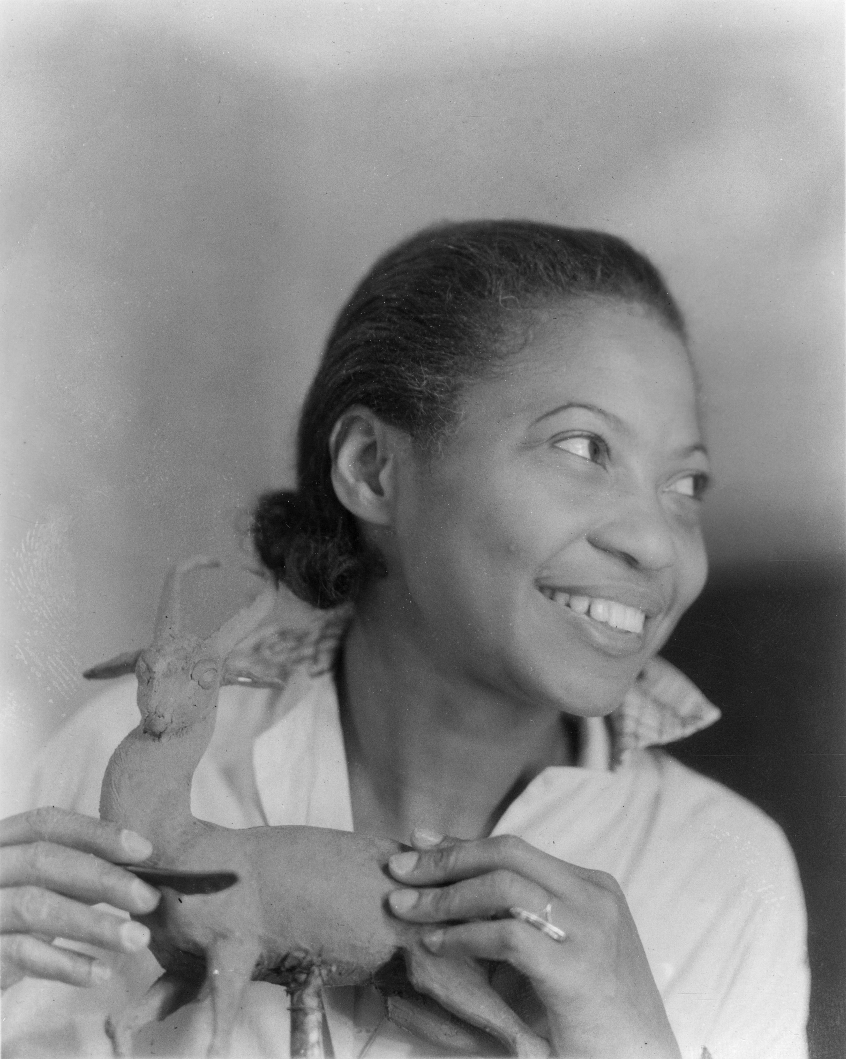 Black and white portrait photo of Augusta Savage. She looks away from the camera to one side with a smile on her face. She is holding a small sculpture. 