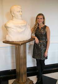 Full color photo of Mallory Mortillaro with her elbow on the stand of a white marble bust of a man. 