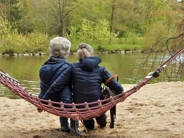 color photo of two women in winter coats sitting in a hammock looking out over a pond