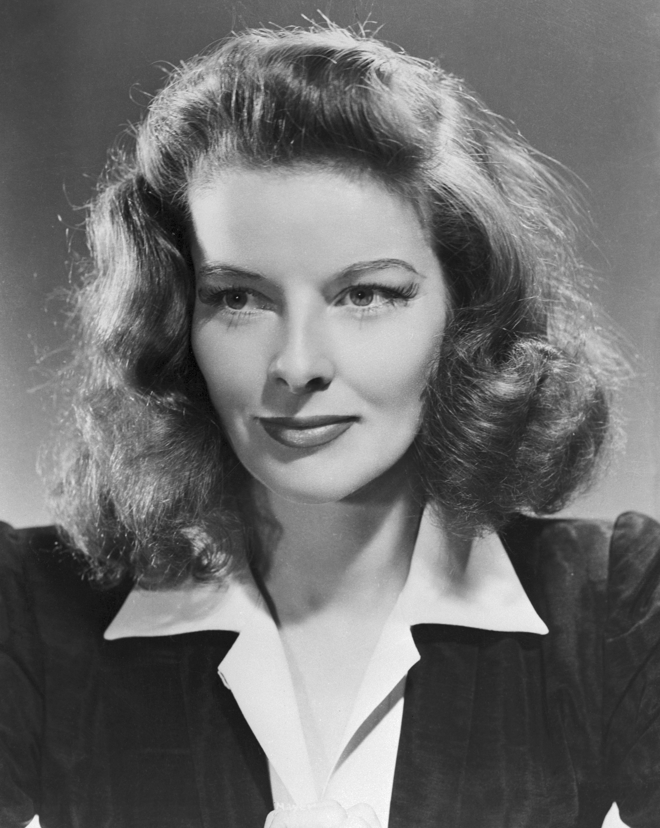 Black and white photo of Katherine Hepburn from the chest up with a small smile, posed as a headshot