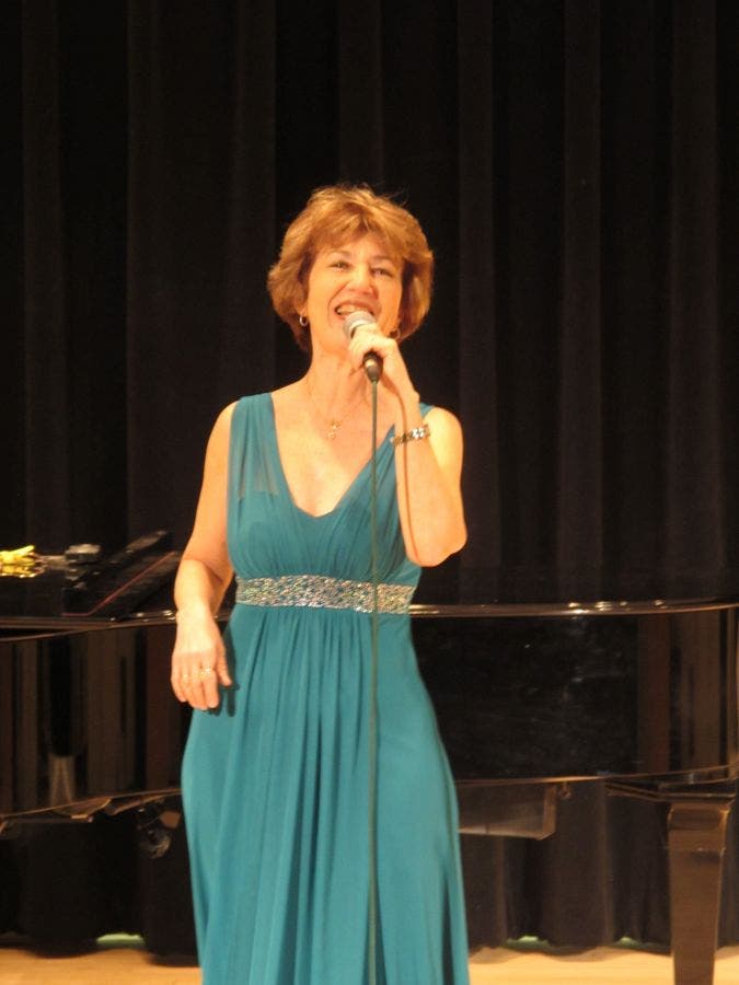 Woman in a teal dress, sparkly belt sings into a microphone
