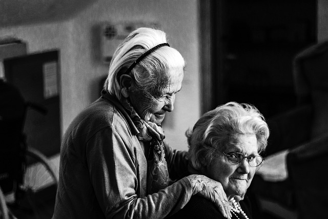 black and white photo of a seated senior woman looking at the camera with another woman standing beside her, hands on her shoulders
