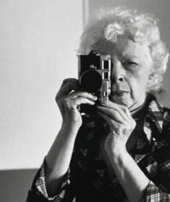 black and white photograph of an woman with white curly hair holding a camera in front of her face 