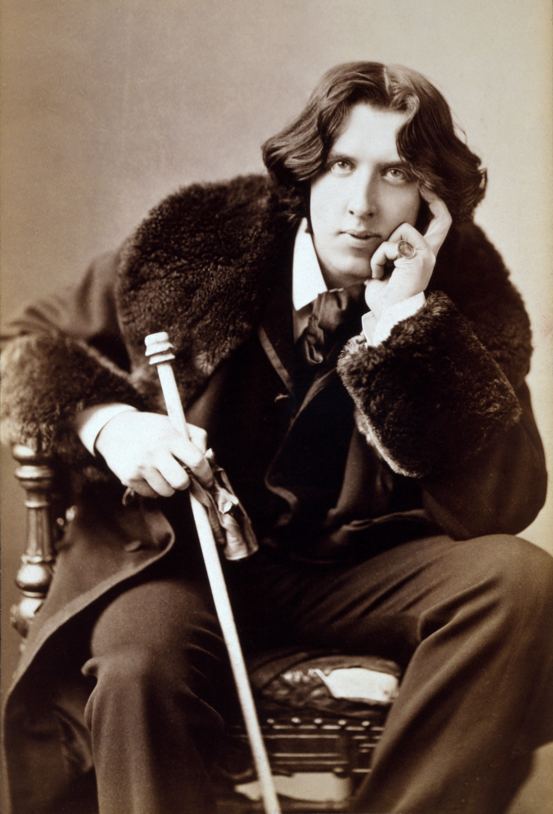 Sepia toned photograph of a young Oscar Wilde in a fur collared coat, holding a cane. He is sitting and his elbow is on his thigh and he cups his face with his hand. 