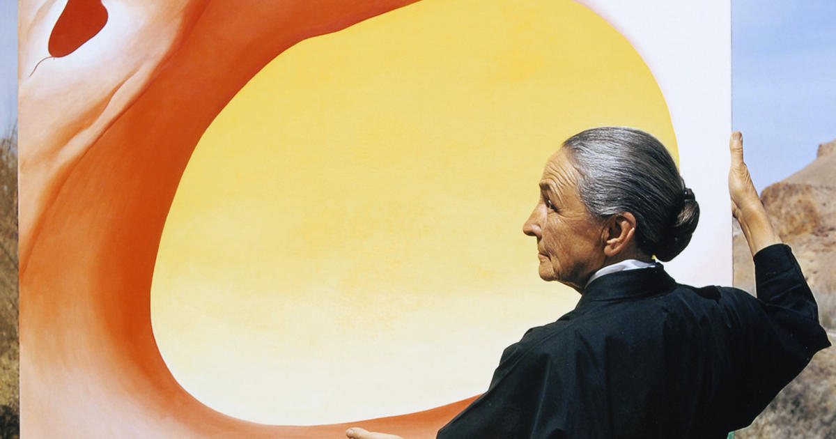 color photo of a woman with grey and white hair in a severe bun, the artist georgia o'keefe, stands in front of one of her works, a circle of yellow surrounded by a red splotch of pain