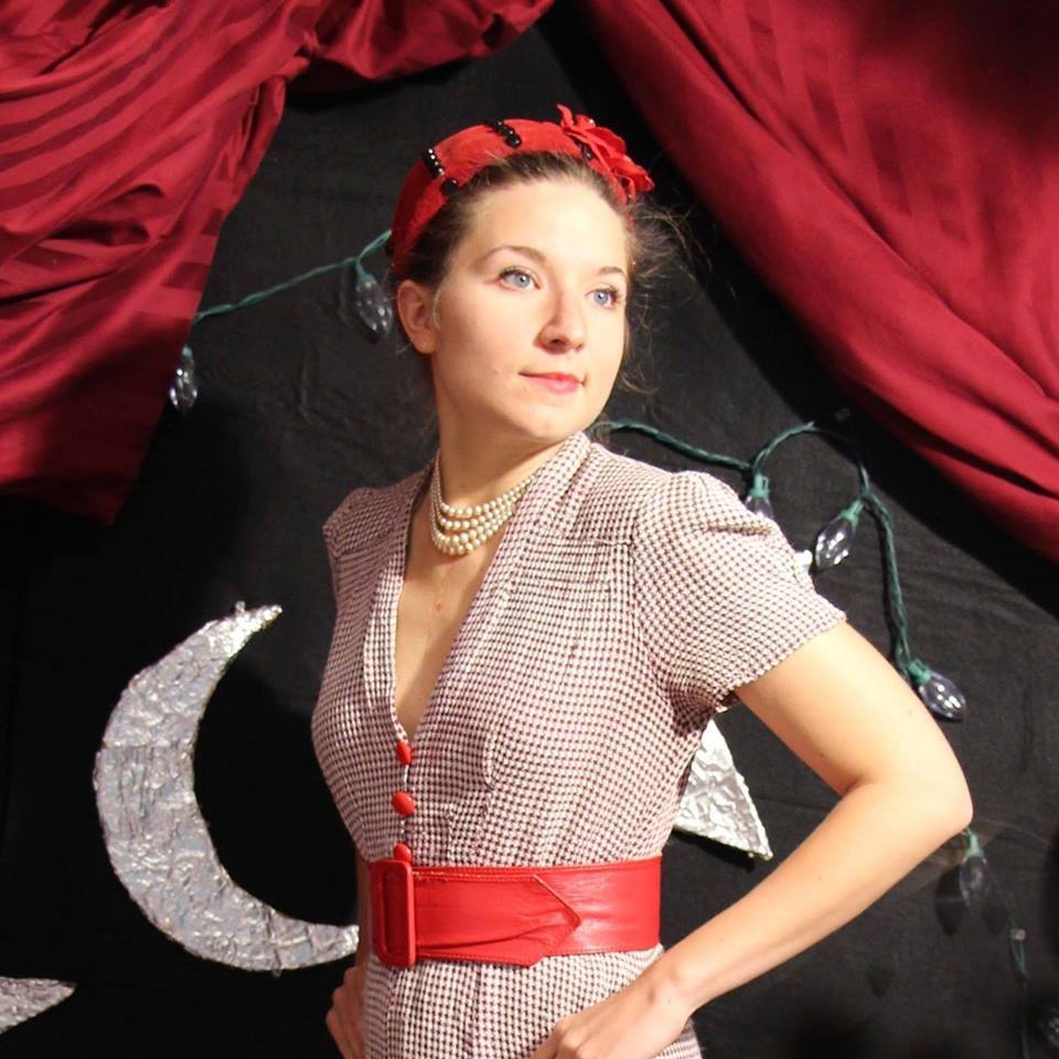 A color photo of Miss Maybell in period dress standing in front of a black curtain with a crescent moon on it. 