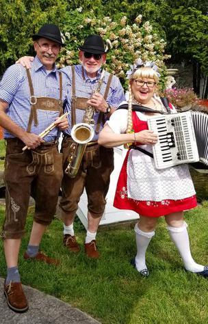 A woman in traditional Swedish clothes holds and accordion in front of two men dressed in lederhosen and wearing caps with feathers. 