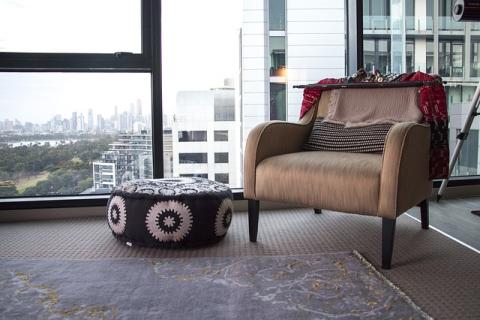 a chair by a large window with a blanket thrown over the back and a decorated round ottoman next to it