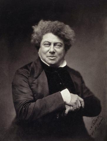 Black and white portrait of Alexander Dumas, he sits in a chair one hand folded over the other and a small smile on his face. 