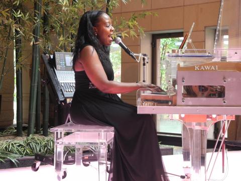 A woman in a long black dress sits at a piano singing into a microphone with a backdrop of indoor greenery and large windows