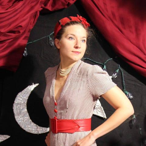 A color photo of Miss Maybell in period dress standing in front of a black curtain with a crescent moon on it. 