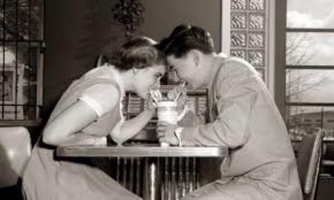 black and white photo of two teenagers in 1950s clothing drinking out of the same milkshake