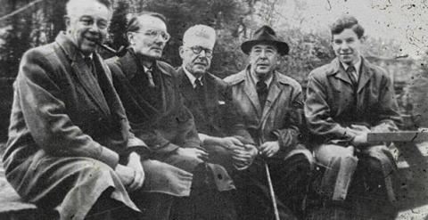 Black and white photo of five men seated on a bench facing the camera. 