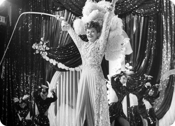 Black and white photo of a woman in a white sparkling neck to ankle dress with an enormous white feathered headpiece