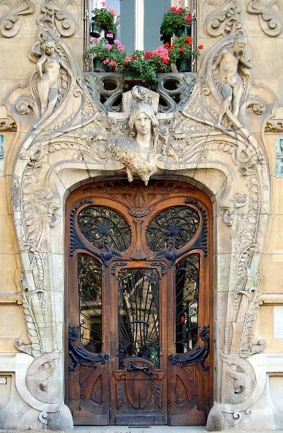 A stone archway over a wooden door. Both are heavily decorated and carved with ivy, leaves and the face of an angel over the door. 